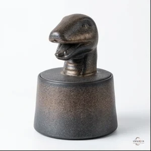 A handcrafted stoneware cremation urn shaped like a snake's head on a cylindrical base, with a speckled dark bronze finish.