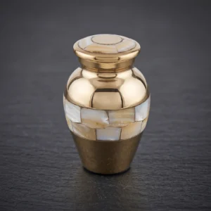 A polished brass mini keepsake cremation urn featuring mother-of-pearl mosaic bands on the body and lid, resting on a dark, textured slate surface.